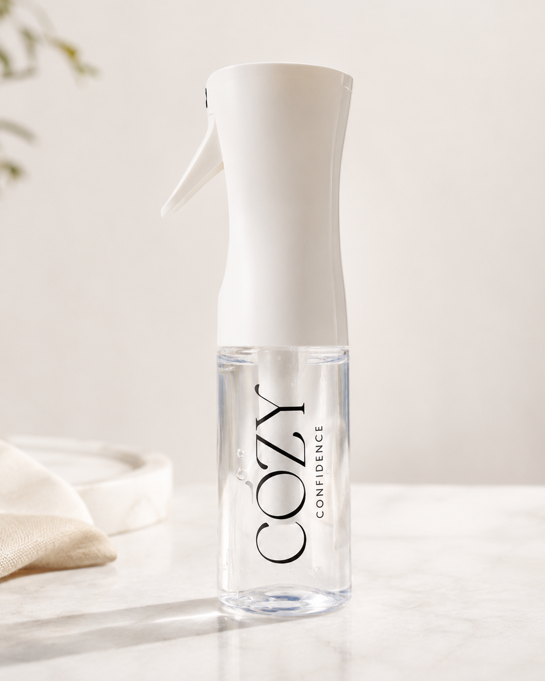 A clear spray bottle with a white trigger top and black 'COZY CONFIDENCE' branding, standing on a light marble surface against a soft cream background.
