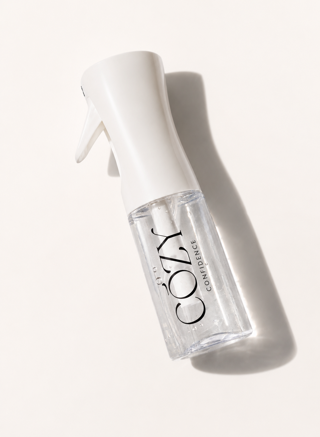 A clear spray bottle with a white spray top and trigger, featuring the brand name 'COZY CONFIDENCE' printed in black on the bottle. The bottle is photographed diagonally on a warm off-white background with a soft shadow cast to the right.