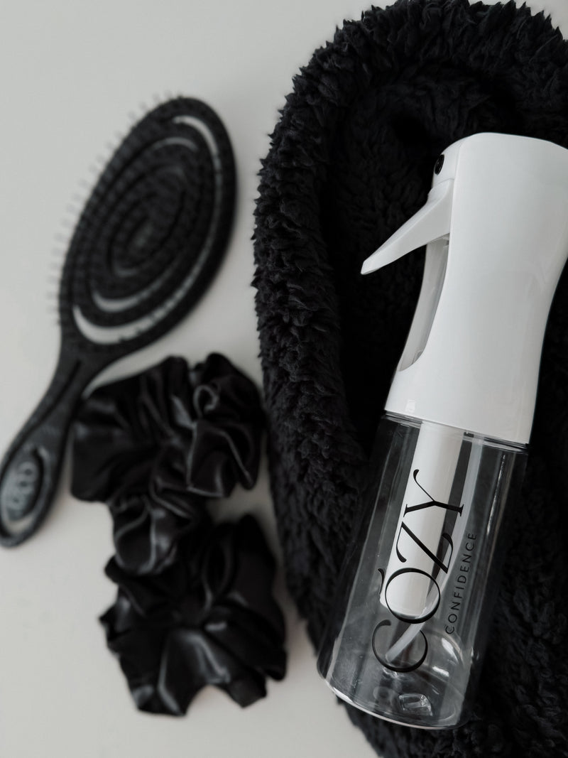 Black Curl Kit flat lay for heatless curls with a rolled black fluffy CurlBand, black silk scrunchies, black Volume Brush and white Cozy Confidence spray bottle