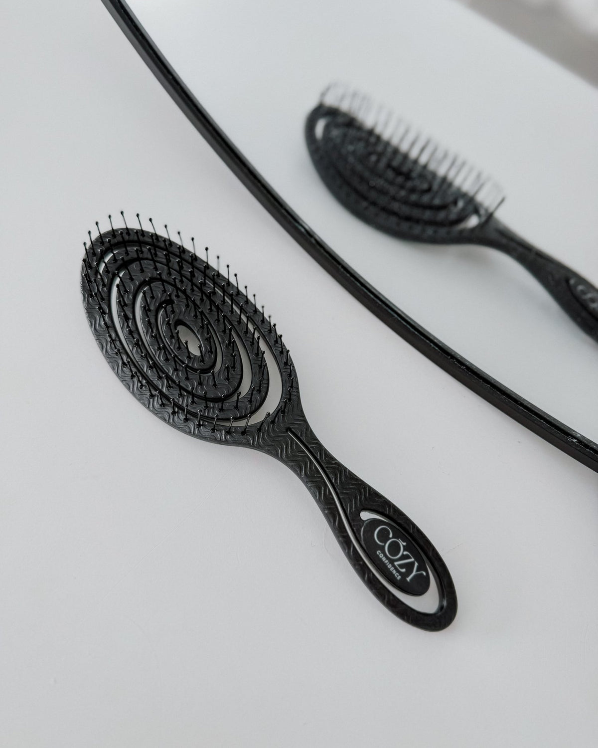Black Cozy Confidence Volume Brush with a spiral vent design and flexible bristles for smoothing heatless curls, shown beside a round mirror