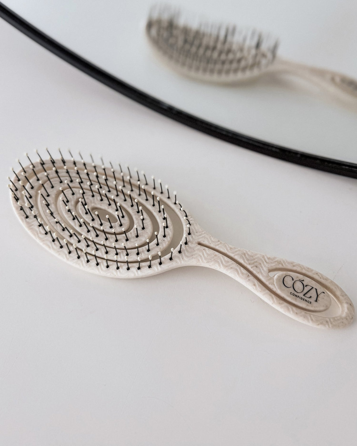 Oatmeal Cozy Confidence Volume Brush with a spiral vent design and black bristles for smoothing heatless curls, reflected in a round mirror