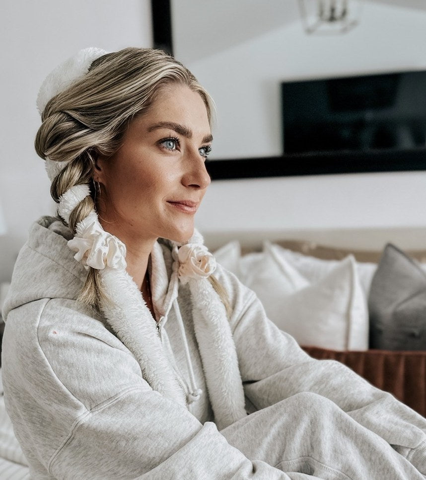 Smiling blonde woman lounging on her bed in a cozy grey sweatsuit, her hair wrapped in a cream CurlBand Blowout with ivory silk scrunchies for heatless curls overnight