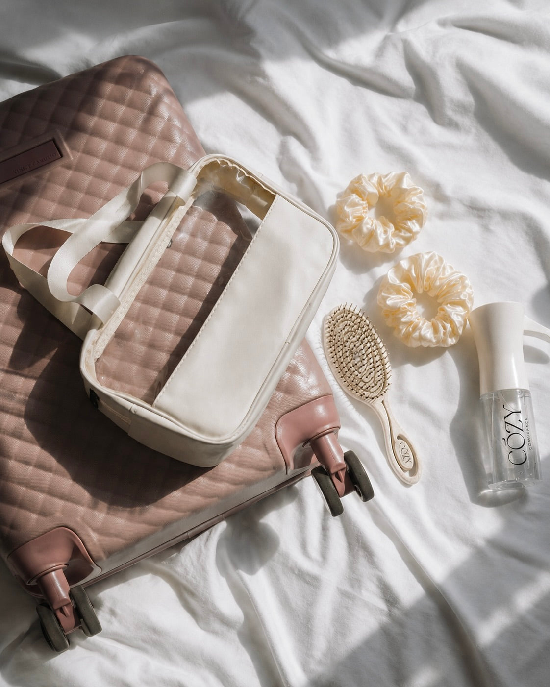 A quilted blush pink suitcase with a cream zippered organizer bag placed on top, styled on white fabric with a Cozy Volume hairbrush, two cream scrunchies, and a small clear Spray bottle labeled COZY.