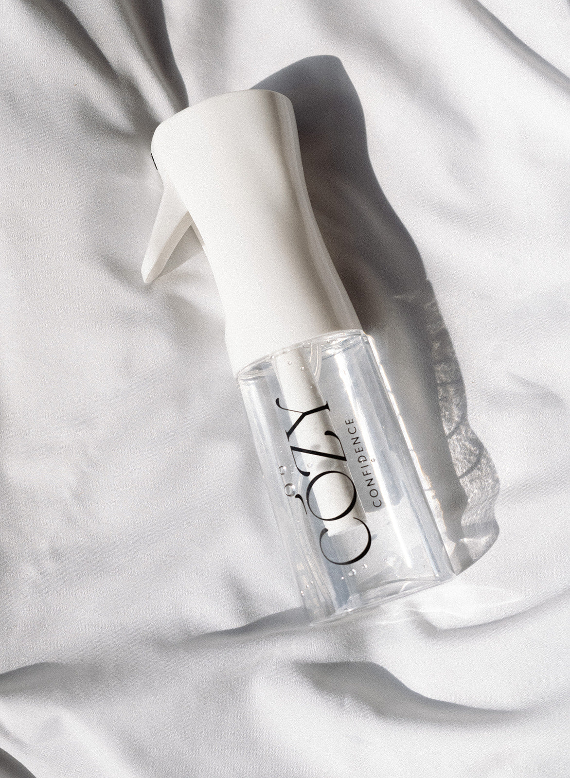 A clear spray bottle with a white trigger sprayer and black 'COZY' branding, resting on draped white fabric with soft shadows.