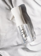 A clear spray bottle with a white trigger sprayer and black 'COZY' branding, resting on draped white fabric with soft shadows.