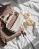 A quilted blush pink suitcase with a cream zippered organizer bag placed on top, styled on white fabric with a Cozy Volume hairbrush, two cream scrunchies, and a small clear Spray bottle labeled COZY.