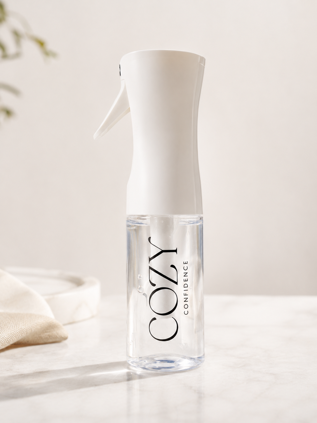 A clear spray bottle with a white trigger top and black 'COZY CONFIDENCE' branding, standing on a light marble surface against a soft cream background.