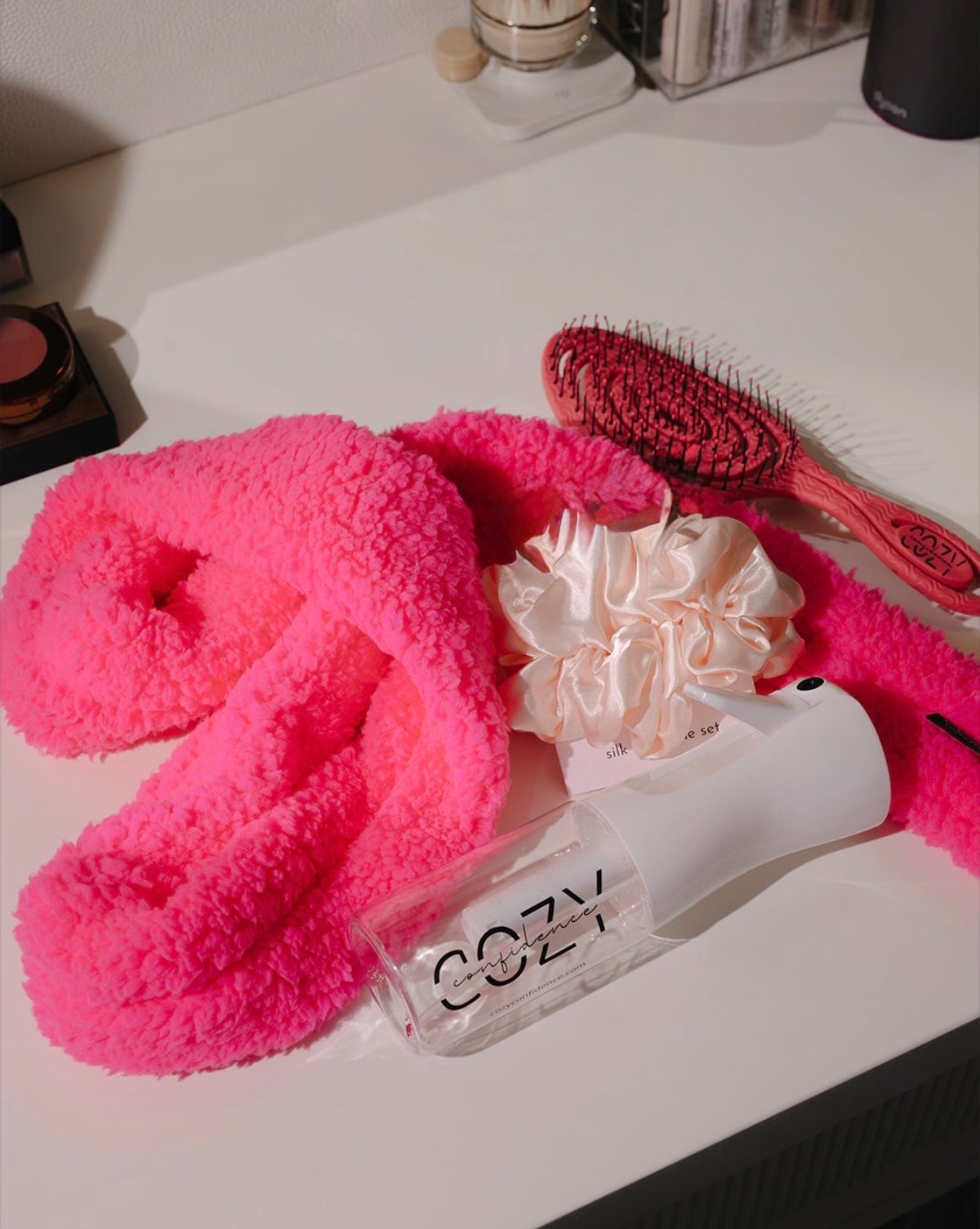Pink CurlBand, satin scrunchie, pink Cozy brush and Cozy spray bottle on a white vanity